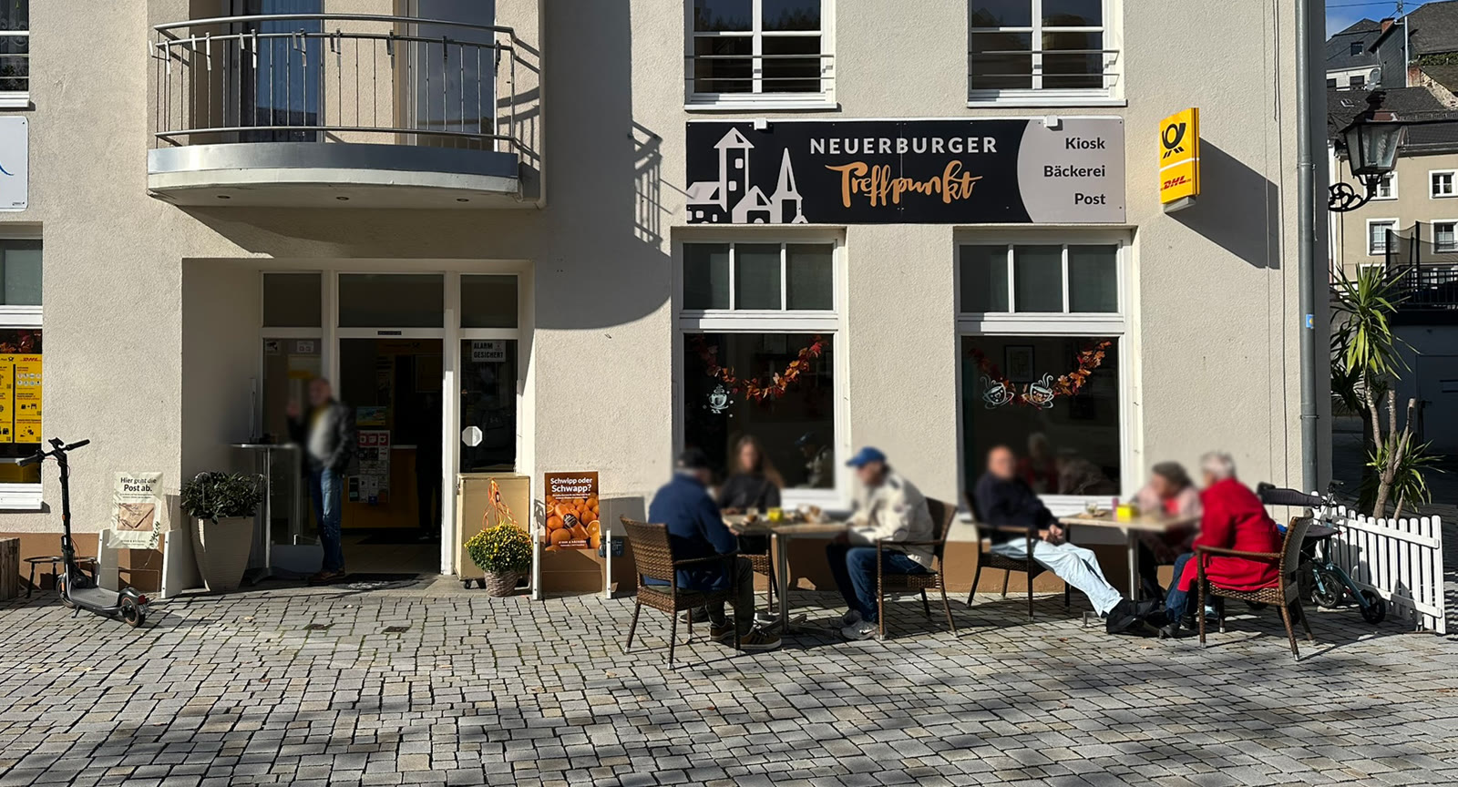 Außenansicht des Neuerburger Treffpunkts mit Logo-Schild, Gäste sitzen draußen in der Sonne